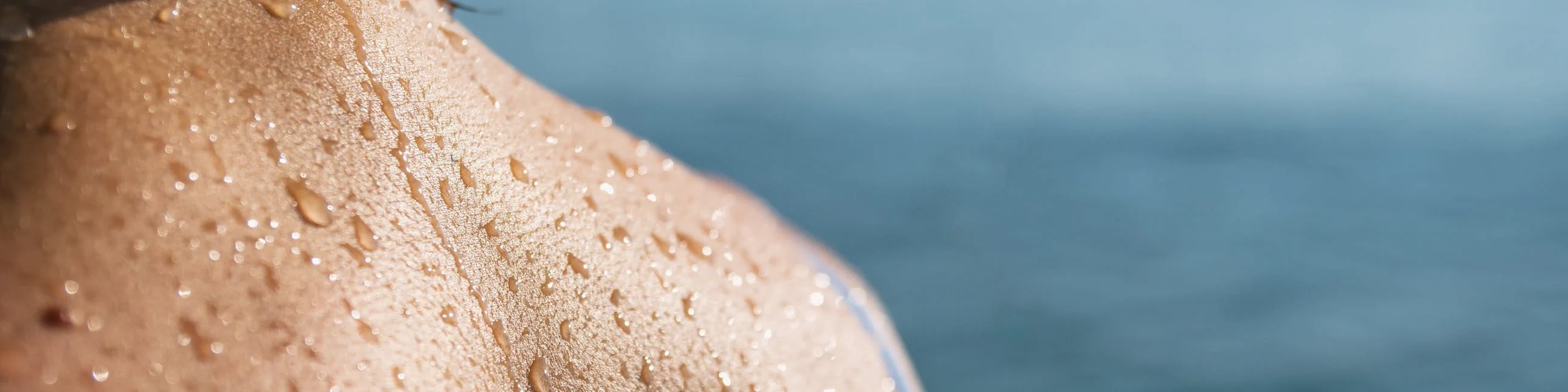 Close-up of a person’s shoulder and upper back with water droplets on the skin, with the sea softly blurred in the background.